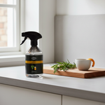 A bottle of Pine & Cedar room spray by The Home Depot stands on a kitchen counter next to a small white cup with a citrus peel and fresh rosemary sprigs on a wooden cutting board.