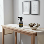 A clear spray bottle with a black trigger top and a black label sits on a light wood table in a bright room. The label has a yellow band and small fruit graphics, but the brand text is not legible.