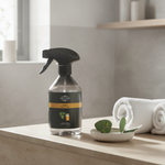 A clear spray bottle of Nature's Way Foot & Skin Cleanser with a black trigger sprayer and black label, set on a light stone countertop next to rolled white towels and a small dish with green leaves, in a softly lit bathroom setting.
