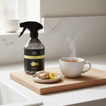A bottle of Ginger Brew cleansing spray stands on a wooden cutting board beside a white mug of steaming tea and ginger slices in a bright kitchen setting.