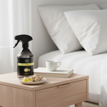 A dark amber spray bottle with a black trigger sprayer and a yellow label sits on a light wood bedside table next to a bed with white bedding. A white mug, a book, and a small plate with ginger and lemon slices are arranged on the table.