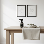 A glass spray bottle with a black trigger sprayer and a black label stands on a light wood table next to a white cloth and a bowl of smooth stones. Two black-framed minimalist line art prints hang on a white wall above the table.
