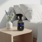 A dark plastic spray bottle with a black trigger sprayer and a purple label band sits on a light wood bedside table next to a white vase with flowers, with a white sofa in the background.