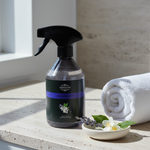 A bottle of Scrutneys Lavender & Jasmine cleaning spray stands on a light stone surface beside a rolled white towel and a small dish with lavender and white flowers. The scene is lit by natural sunlight with soft shadows.