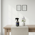 A dark glass spray bottle with a black trigger sprayer and a purple label sits on a light wood table runner next to a small white vase with white and purple flowers. The background is a clean white wall with two minimalist black-framed art prints.