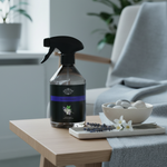 A spray bottle of Kenwood Home Refreshing Air Freshener sits on a light wood side table in a cozy living room setting, next to a bowl of bath stones and white flowers.