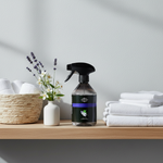 A spray bottle of cleaning product with a black trigger sprayer and dark label stands on a wooden shelf beside a woven basket, a small white vase with flowers, and folded white towels against a light gray wall.