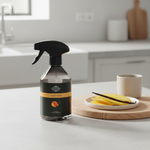 A clear spray bottle with a black trigger sprayer and a black-and-gold label reading 'Scent of Home' and 'Vanilla & Mango' sits on a light countertop in a bright kitchen. A small plate with yellow fruit slices and a white cup are on a wooden board beside it.