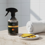 A bottle of Clorox Vanilla & Mango cleaning spray stands on a stone countertop beside a rolled white towel and a small plate with vanilla beans, against a tiled wall.