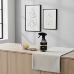 A glass spray bottle with a black trigger sprayer and black label sits on a light wood countertop beside a white towel, with framed abstract wall art in the background.