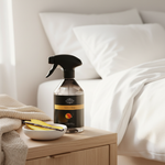 A Kellie Rose 'Natural Shine' glass spray bottle with a black trigger sprayer and black label, styled on a light wood bedside table next to a bed with white linens.