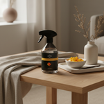 A clear glass spray bottle with a black trigger sprayer and a black label featuring a yellow band and a small orange fruit graphic, set on a light wood table in a softly lit home interior.