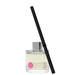 A clear glass diffuser bottle with a pink label and a black reed stick, containing a yellow liquid.